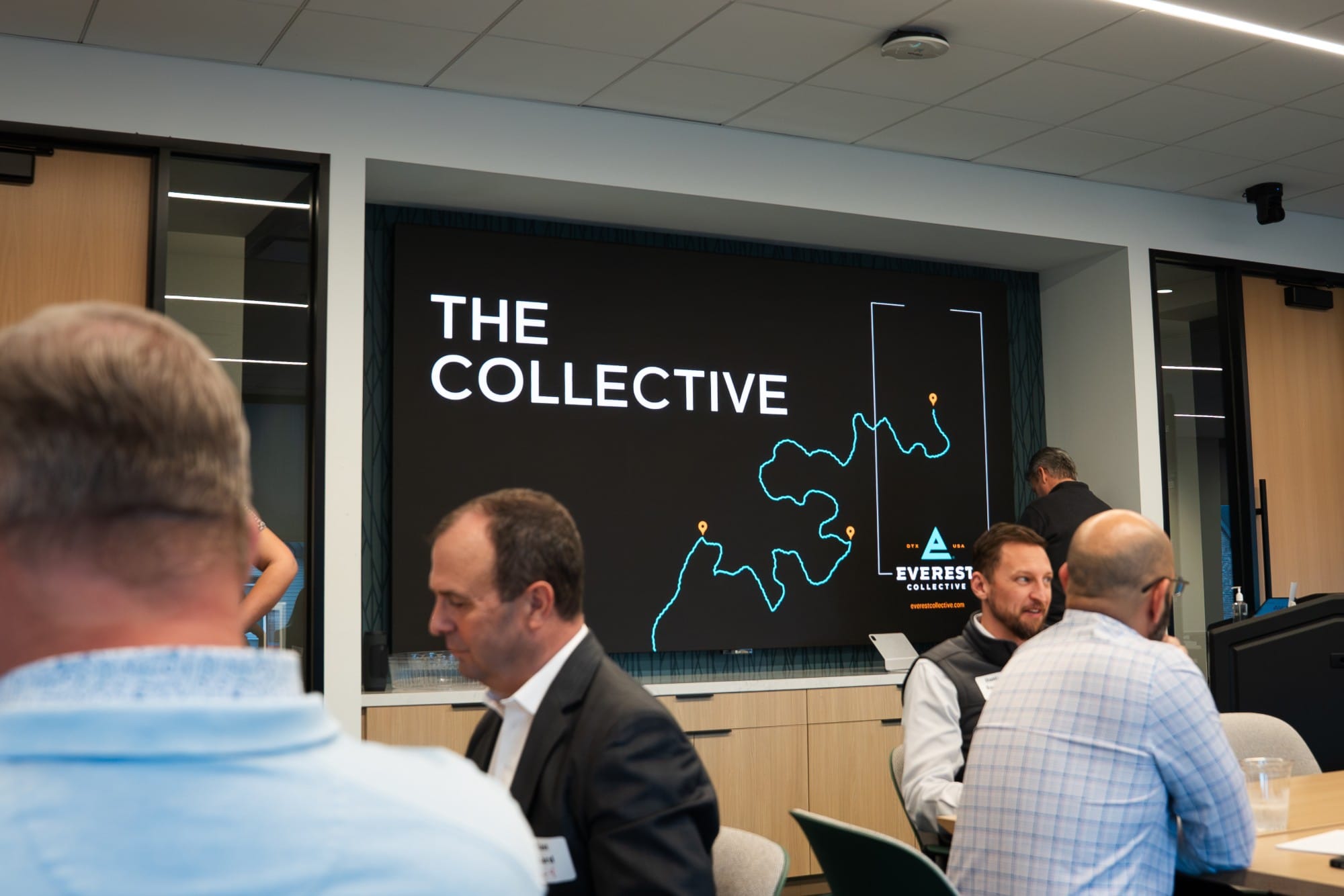 Executives at tables in front of a screen that reads The Collective