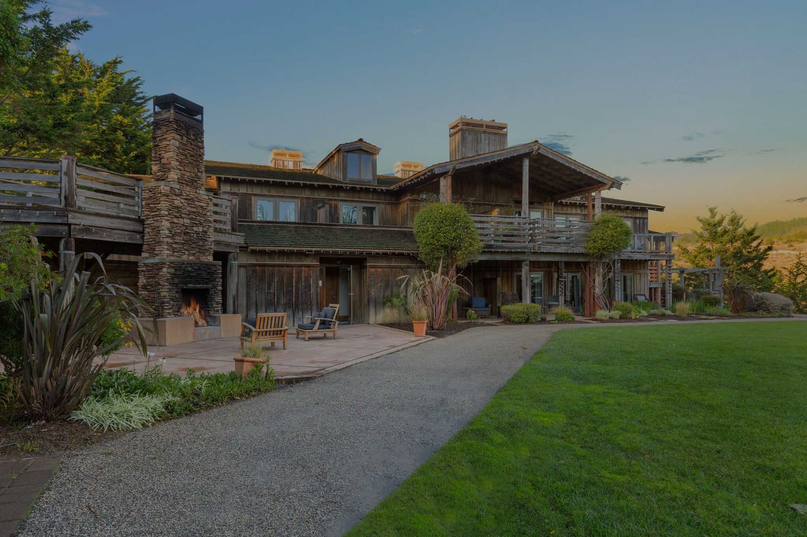 Weathered wood lodge with a stone outdoor fireplace and lawn at dusk