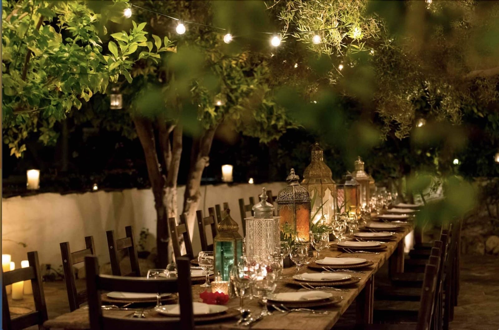 A long candlelit dinner table under string lights and trees
