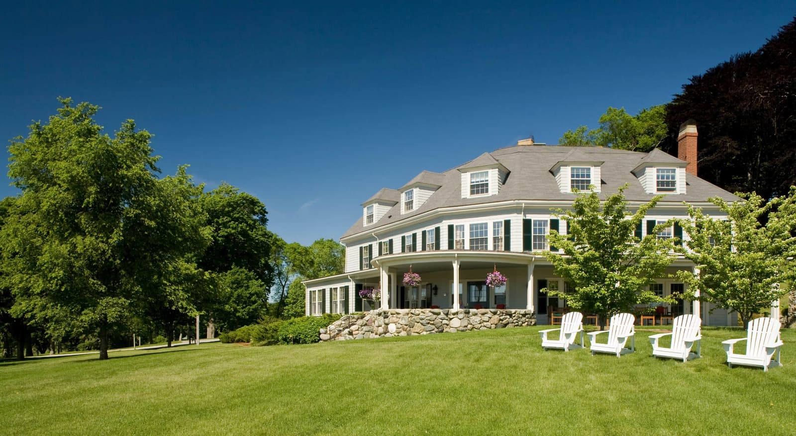 The white shingled inn with a wraparound porch and Adirondack chairs on a green lawn