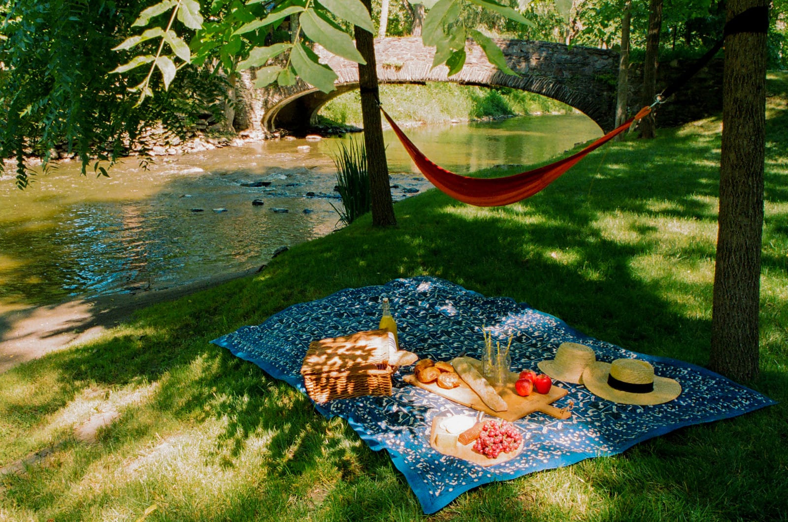 A picnic blanket and a red hammock beside a stream under shade trees