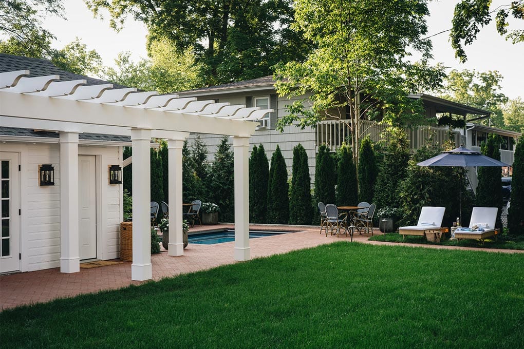 A white pergola and patio beside a green lawn and tall hedges